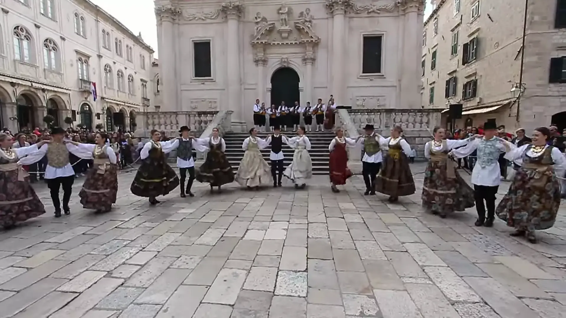 Dancers in dark embroidered skirts, white blouses and waistcoats form a line across a stone-paved Dubrovnik square, with an orchestra on the steps of a baroque church behind them.