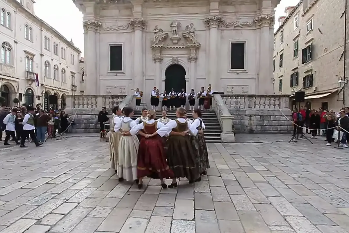 Women in long full skirts and white blouses form a tight circle on stone paving while an orchestra stands on the steps of a baroque church.