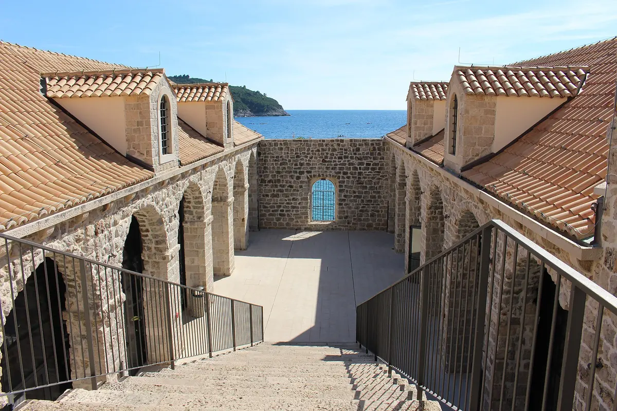 Stone arcades and terracotta roofs around a courtyard, with a stairway leading down and the sea visible through an opening at the far end.