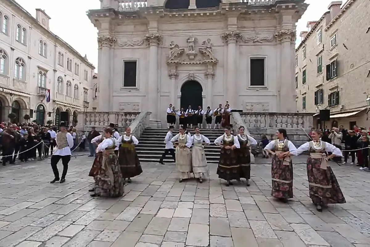 A line of dancers in costume, men in black hats and women in long skirts, stands across the paved square in front of the church steps where the orchestra plays.