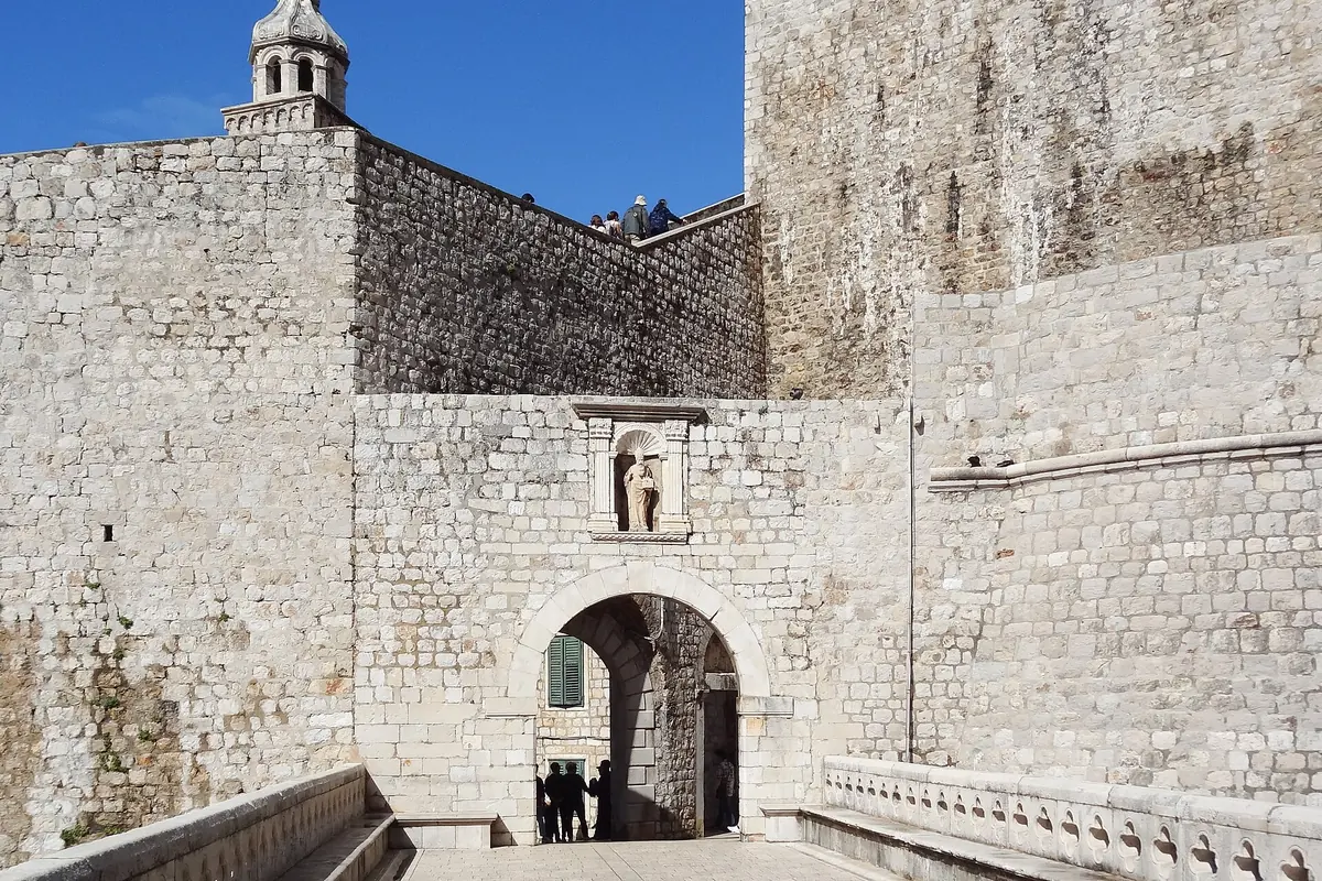Ploče Gate in Dubrovnik's stone walls, with a stone bridge leading to an arched entrance and a statue in a niche above it